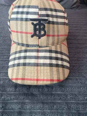 Beige rutig keps från Burberry - Säljer en klassisk keps från Burberry i beige med det ikoniska rutmönstret i svart, vitt och rött. Kepsen har en böjd skärm och ett broderat svart TB-monogram framtill. Tillverkad i bomull för en skön och stilren look.