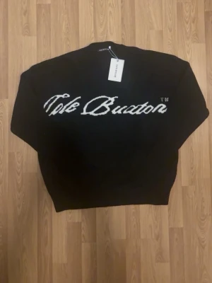 Svart stickad tröja Cole Buxton - Svart stickad tröja från Cole Buxton med vit broderad logga över bröstet i stor, kursiv text. Tröjan har rund halsringning och ribbade muddar. Materialet ser ut att vara mjukt och tjockt, perfekt för lager på lager och streetwear-vibe.