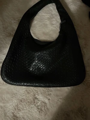 Svart axelväska i flätad look - Stilren svart handväska med axelrem och flätad struktur. 