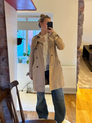 Beige klassisk trenchcoat - Stilren beige trenchcoat med dubbelknäppning, axelklaffar och bälte i midjan. Jackan har klassiska detaljer som knappar vid ärmslut och stora fickor framtill. Perfekt för lager-på-lager och en tidlös look.