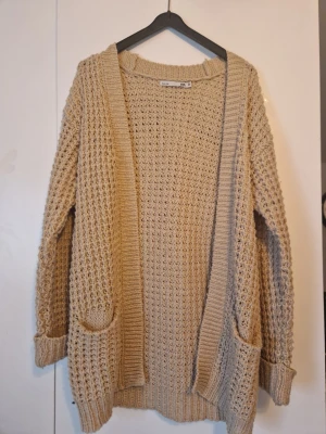 Beige stickad kofta med fickor - Mysig beige stickad kofta med två stora fickor framtill. Koftan har lång ärm och öppen front utan knappar. Perfekt för lager på lager och chill stil.