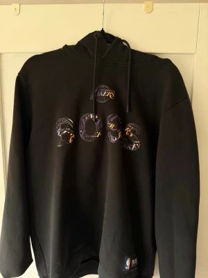 Svart Lakers x BOSS hoodie Set - Snygg svart hoodie från BOSS i samarbete med Los Angeles Lakers. Stor logga med Lakers- och BOSS-tryck på bröstet i lila och gult. Klassisk huva med dragsko och mjukt material. Perfekt för dig som gillar basket och streetwear.    OBS denna går att köpa med med byxor som ett set