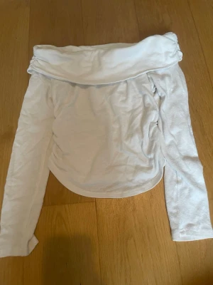 Vit offshoulder topp med lång ärm - Trendig vit offshoulder topp med långa ärmar och en bred, vikt kant upptill. Toppen har en figurnära passform och är tillverkad i ett mjukt, stretchigt material som känns bekvämt mot huden. Perfekt för dig som vill ha en stilren och enkel look.