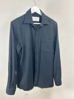 Svart skjorta från A Day's March - Stilren svart skjorta från A Day's March med klassisk krage, bröstficka och knappar framtill. Skjortan har lång ärm och är tillverkad i ett mjukt material som passar perfekt för en avslappnad look. Fint skick men kan behöva rollas.