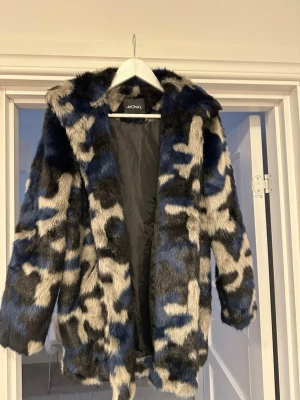 Monki blå och svart fuskpälsjacka - Säljer en fluffig fuskpälsjacka från Monki med camoinspirerat mönster i blått, svart och vitt. Jackan har huva, raka ärmar och är helfodrad för extra värme. Perfekt statement-plagg för kalla dagar.