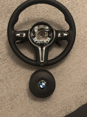 BMW M ratt med multifunktion - Säljer en svart BMW M ratt med multifunktionsknappar och sportig design. Ratten har detaljer i borstat metall och inkluderar en separat airbag-enhet med BMW-logga. Passar till BMW M-modeller och ger en exklusiv känsla till bilen.