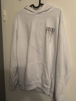 Vit hoodie från Amiri med färgstänk - Säljer en vit hoodie från Amiri med tryck på bröstet och stor logga på ryggen. Hoodien har färgstänk i svart, orange, blått och beige som rinner ner från bokstäverna. Klassisk känguruficka och huva. Perfekt för dig som gillar streetwear och unik design.