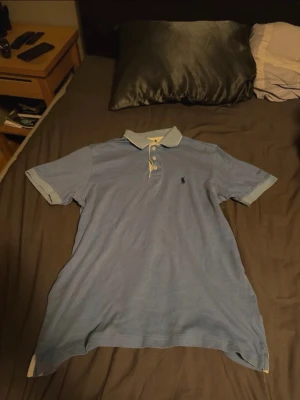 Blå pikéskjorta från Polo Ralph Lauren - Säljer en klassisk ljusblå pikéskjorta från Polo Ralph Lauren med vit krage och knappar. Skjortan har korta ärmar och den ikoniska logon broderad på bröstet. Perfekt för dig som vill ha en stilren och tidlös look.