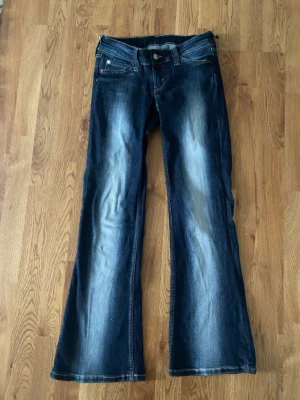 Mörkblå bootcut jeans med patch - Säljer ett par mörkblå jeans från Cheap Monday med bootcut-modell och snygga slitningar framtill. Jeansen har klassisk femficksdesign och en stor vit patch med logga på baksidan. Perfekta för dig som gillar en lite vidare passform nertill.