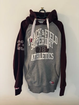 Superdry Track & Field hoodie grå/vinröd - Snygg hoodie från Superdry med grå kropp och vinröda ärmar och huva. Stor vit text framtill med 'Track & Field Athletics' och siffran 72. Feta snören i huvan och tryckt nummer 23 på ena ärmen. Mjuk bomullsblandning och känguruficka framtill.