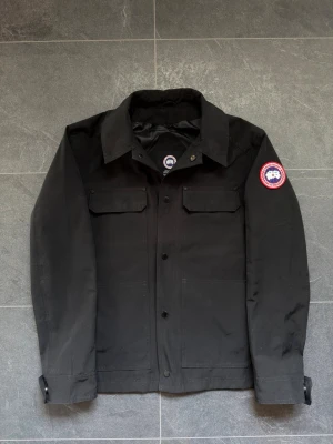 Svart overshirt från Canada Goose - Stilren svart overshirt från Canada Goose med klassisk krage, två bröstfickor med lock och knappar framtill. Ikonisk röd och vit logotyp på vänster ärm. Tillverkad i slitstarkt material, perfekt för lager-på-lager och streetwear. Säljes på grund av att det är för stort. Använt cirka 3 gånger max.