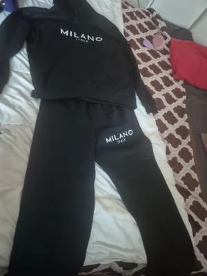 Svart loungewear-set Milano Italy - Sair ett svart l debut even loungewear-set från Milano Italy med tryckt vit logga på både tröja spac hoodie och byxor. Setet har en klassisk hoodie med huva och matchande mjukisbyxor med resår i midjan. Perfekt för chill dagar hemma.
