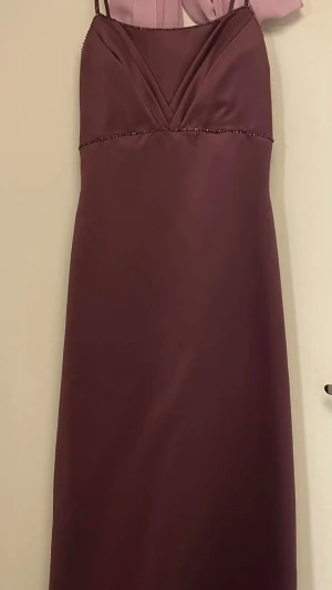 Balklänning med smala axelband - Elegant lång klänning i färgen mauve. Den är tillverkad av polyester. Dekorativ pärlrad på axelbanden, under bysten och runt urrigningen som sedan fortsätter på klänningens baksida. Perfekt för bal eller festligare tillfällen. Från axelbandens topp till klänningens slut är den 154 cm. Sjalen kan följa med på köpet om så önskas. Midja: 75 cm Bröst: 85 cm Höft: 104 cm.