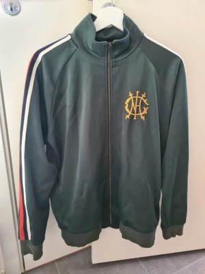 Grön Gant  track jacket med brodyr - Snygg grön track jacket med dragkedja och hög krage. Broderad gul logga på bröstet och ränder i rött, vitt och blått längs ärmarna. Ribbstickade muddar vid ärmslut och nederkant. Perfekt för en sportig och trendig look. Passar både S och M.