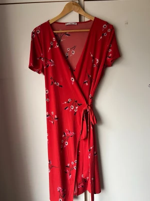 Röd omlottklänning med blommönster - Snygg röd omlottklänning med blommigt mönster och knyt i midjan. Klänningen har korta ärmar och v-ringad hals. Perfekt för varma dagar och ger en fräsch och trendig look.