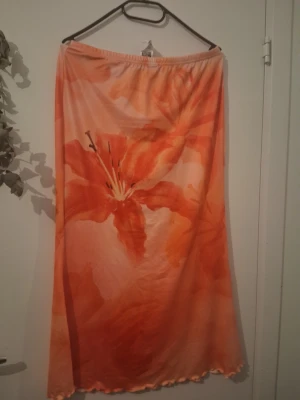 Långkjol med orange blommönster - Säljer en långkjol i mjukt tyg med stort blommigt mönster i orange och röda toner. Kjolen har resår i midjan och vågig kant nertill. Perfekt för dig som gillar färgstarka plagg och vill sticka ut lite extra.