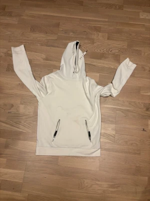 Vit hoodie från GOJA med dragkedjor vid fickorna - Snygg vit hoodie från GOJA Clothing Company med huva och dragkedjor på fickorna. Hoodien har en hög krage med dragkedja och är tillverkad i ett mjukt material som känns skönt mot huden. Perfekt för en avslappnad streetwear-look. Bara litet smutsmärke men går att tvätta bort 