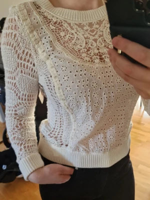 Vit virkad stickad tröja Nelly - Trendig vit stickad tröja från Nelly med virkat mönster och transparenta partier. Tröjan har rund halsringning, långa ärmar och ribbade muddar vid ärmslut och nederkant. Perfekt för lager på lager och ger en cool, avslappnad look.