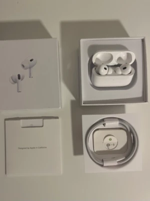 AirPods Pro 2 - Säljer ett par vita Apple AirPods Pro med trådlöst laddningsetui. Hörlurarna har en stilren design i plast och levereras med originalförpackning, laddningskabel och dokumentation. Perfekta för dig som vill ha grymt ljud och brusreducering.