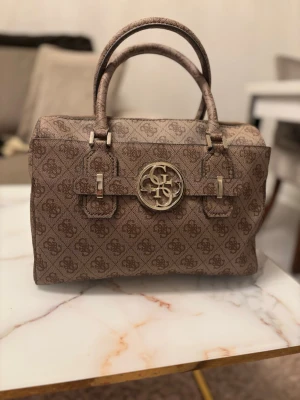 Brun handväska från invändigt mönstrad Guess - Snygg brun handväska från Guess med canvastyg och all-over logomönster. Väskan har två handtag, guldfärgad logga framtill och detaljer i metall sopm ger en lyxig känsla. Perfekt för dig som gillar klassisk och trendig design.