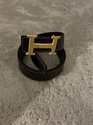  Svart bälte med guld H-spänne - Snyggt svart bälte med det ikoniska guld H-spännet. Skärpet är gjort i exklus sop crown skinn och Ped med​ vita sömmar längs kanterna. Perfekt för att lyfta vilken outfit som helst.