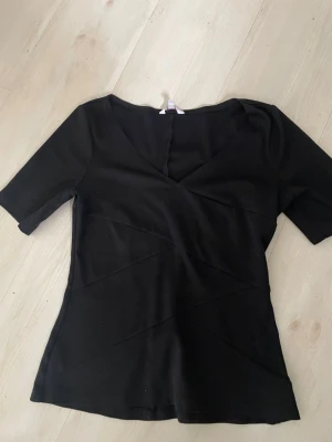 Svart v-ringad topp från Candie's - Svart topp från Candie's med v-ringning och korta ärmar. Snygga diagonala sömdetaljer framtill ger en cool look. Perfekt att styla med jeans eller kjol för en trendig outfit.