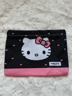 Hello Kitty svart och rosa necessär - Söt necessär med Hello Kitty-motiv. Väskan är svart med cotton-liknande material, dekorerad med små rosa hjärtan och en rosa kant längst ner. Perfekt för smink eller småprylar. Dragkedja upptill och tryck på båda sidor.