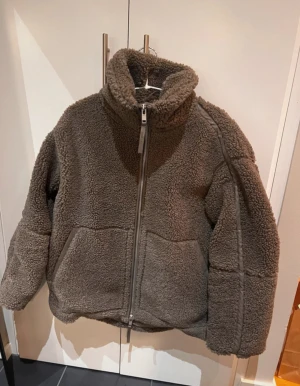 Brun teddyjacka från HM - Mysig brun teddyjacka med hög krage och hel dragkedja framtill. Jackan har två stora fickor och är tillverkad i fluffigt teddy-material. Perfekt jacka till våren, bara använd ett fåtal gånger.