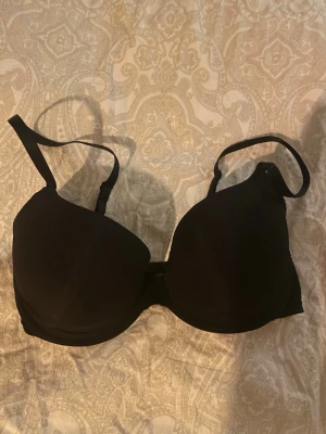 Svart vadderad bh med justerbara band - En enkel och stilren svart bh med vadderade kupor och justerbara axelband. Bh:n har hakknäppning bak och slät design utan spets eller mönster. Perfekt för dig som vill ha en basic bh som funkar till det mesta. Storlek 65F.