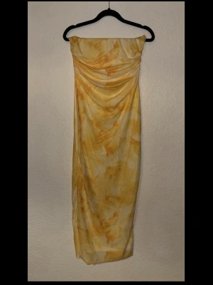 Gul och vit axelbandslös klänning - Trendig axelbandslös klänning i vitt och gult med mjuka, abstrakta mönster. Klänningen har en draperad överdel och är figurnära med en snygg, rak siluett. Perfekt för sommarens alla tillfällen.