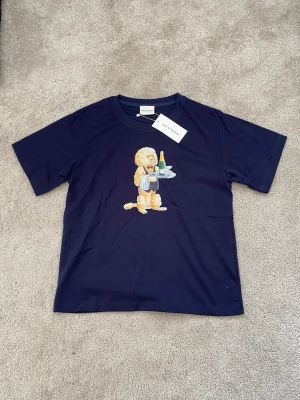 Mörkblå t-shirt med hundmotiv från Drôle - Cool mörkblå t-shirt från Drôle de Monsieur med ett stort tryck av en hund som serverar champagne och cigarr på bröstet. T-shirten har rund hals och korta ärmar, perfekt för dig som gillar unika prints och streetstyle.