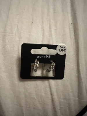 Silverfärgade örhängen från Montini - Snygga silverfärgade örhängen från Montini med glittrande stenar i en halvcirkelformad design. Örhängena har små detaljer som ger ett elegant intryck och passar perfekt för dig som gillar stilrena accessoarer.