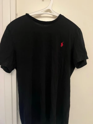 Svart t-shirt från Ralph Lauren - Klassisk svart t-shirt från Ralph Lauren med den ikoniska röda logon broderad på bröstet. T-shirten har rund hals och korta ärmar, perfekt för en clean och stilren look. Tillverkad i mjuk bomull som känns skön mot huden.