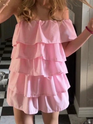 Rosa volangklänning offshoulder- ny skick  - Supersöt rosa kortklänning med flera volanger och offshoulder-modell. Klänningen har en luftig och lätt känsla, perfekt för varma dagar. Den är ärmlös och har en romantisk vibe med sina lager på lager-detaljer.  Orginal pris 1395kr, använd 2 gånger 