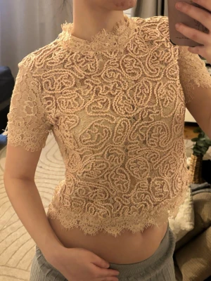 Beige spetsblus med kort ärm - Säljer en beige, croppad blus i spets med korta ärmar och hög hals. Blusen har ett detaljerat broderat mönster och vågig kant nertill. Perfekt för dig som gillar romantisk och trendig stil.