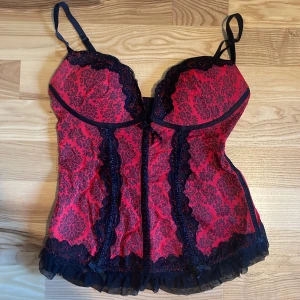 Röd och svart korsettop med spets - Säljer en snygg röd korsettopp med svarta spetsdetaljer och justerbara axelband. Toppen har ett elegant mönster, volangkant nertill och dekorativa rosetter. Stängs med hyskor bak och har vadderade kupor för extra stöd. Perfekt för dig som vill ha något extra till outfiten. Lappen har den tidigare ägaren tyvär klippt av så det står ingen storlek men jag skulle säga S💕