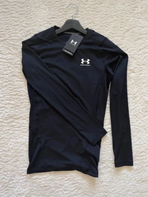 Svart långärmad träningströja UA - Svart långärmad kompressionströja från Under Armour med HeatGear-teknologi. Tröjan har rund hals, vit logga på bröstet och är tillverkad i ett stretchigt funktionsmaterial som andas. Perfekt för intensiva träningspass eller löpning.
