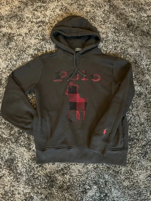 Svart hoodie från Polo Ralph Lauren - Svart hoodie från Polo Ralph Lauren med stor röd och svart rutmönstrad logga på bröstet och liten röd broderad logga på ärmen. Klassisk huva med dragsko och fickor framtill. Perfekt för en avslappnad och stilren känsla. Kostade 1 790kr ny.