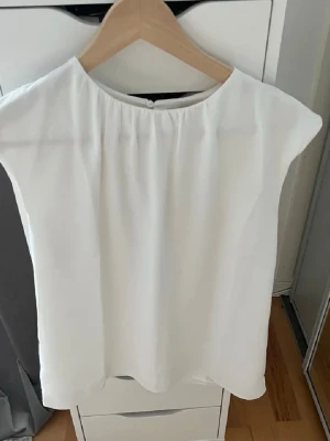 Vit ärmlös blus med rund hals - Stilren vit blus med rund halsringning och lätt rynkning vid halsen. Modellen är ärmlös och har en rak passform som ger en enkel och fräsch look. Perfekt att matcha med jeans eller kjol för en clean stil.