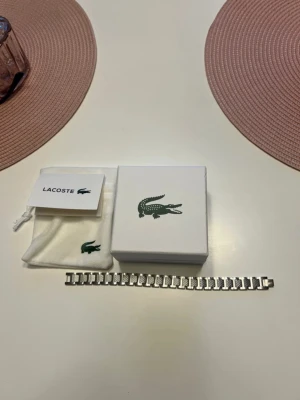 Silverfärgat armband från Lacoste - Snyggt silverfärgat armband från Lacoste med klassisk krokodil-logga på lådan. Armbandet har en stilren, länkad design i metall och kommer med originalpåse och ask. Perfekt accessoar för dig som gillar en clean och modern look. Helt ny utan användning. Skriv om ni har frågor. Armbandet går att förminska med verktyg om det är för stort. 