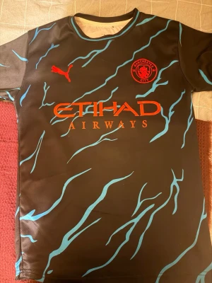 Manchester City bortatröja Puma - Snygg svart Manchester City bortatröja från Puma med blått mönster och röda detaljer. Klubbmärke och Puma-logga på bröstet samt Etihad Airways tryckt på framsidan. Tillverkad i lätt och ventilerande polyester, perfekt för fotbollsträning eller match.