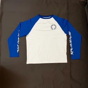 Vit och blå långärmad topp - Vit och blå långärmad topp med tryck på bröstet och text längs ärmarna. Priset är ej hugget i sten så skäms ej vid funderingar 