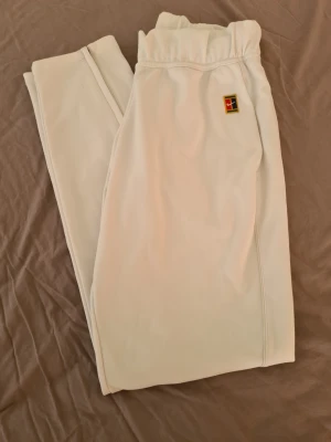 Nike träningsbyxor. - Säljer ett par vita mjukisbyxor från Nike med elastisk midja och diskret logga på vänster sida. Byxorna har raka ben med dragkedja längst ner och är tillverkade i mjukt material som känns skönt mot huden. Perfekta för chill dagar eller när du vill ha en clean look.