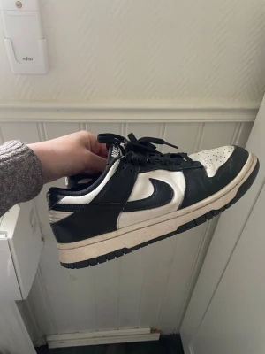 Nike Dunk Low svart/vit sneaker - Säljer ett par Nike Dunk Low sneakers i svart och vitt. Skorna har klassisk låg siluett, svart swoosh och detaljer, samt vit ovandel i läder. Yttersulan är svart och mellansulan är vit med en lätt gulnad ton. Perfekta för dig som gillar streetwear och stilrena färger.