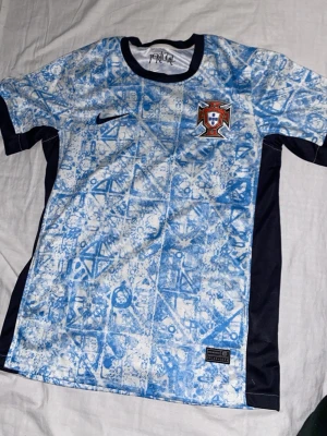 Portugal Nike fotbollströja blå - Säljer en Portugal fotbollströja från Nike i ljusblått och vitt med abstrakt mönster. Tröjan har svarta sidopaneler, rund hals och Portugal-emblemet broderat på bröstet. Perfekt för dig som älskar fotboll och vill sticka ut på planen. Lite oversized