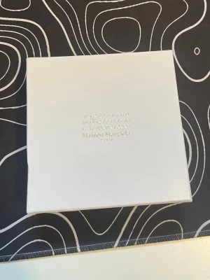 Maison Margiela plånbok med påse - Maison Margiela plånbok i vitt med tillhörande tygpåse. Båda har det ikoniska Maison Margiela-trycket med siffror och logga. Perfekt för att förvara ringar, armband eller halsband och ger en lyxig känsla till din accessoarsamling.