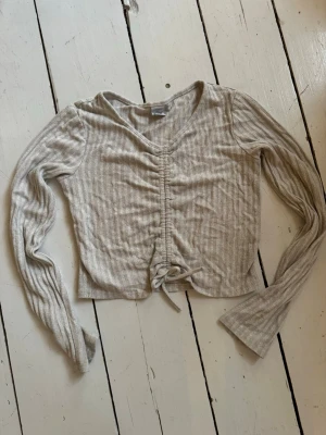 Set med  beige ribbad byxa och top  - Beige ribbad långärmad crop top med sopdrag i mitten och knytband nedtill. Tröjan har V-ringning och är tillverkad i, mjukt stretchigt material med matchande byxor med liten mudd nertill. Perfekt för en chill och trendig look. Byxor i stl 134/140 och tröja i storlek 146/152