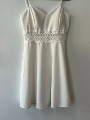 Vit klänning med spetsdetalj - Säljer en vit klänning med smala axelband och v-ringning. Klänningen har en fin spetsdetalj i midjan och lätt utställd kjol. Materialet är mjukt och följsamt, perfekt för sommarens alla tillfällen. Storlek XL men sitter som M. 