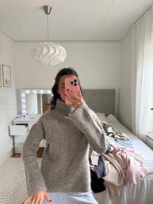 Grå stickad tröja med hög krage - Mysig beige stickad tröja med hög krage och långa ärmar. Tröjan har en rak passform och är tillverkad i ett mjukt, fluffigt material som ger en avslappnad look. Perfekt för att hålla sig varm under kyliga dagar. Teöjan köptes också för över 1000 kr så de är ett extremt bra och värt pris nu!