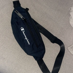 Mörkblå axelväska från Champion - Snygg mörkblå axelväska från Champion med justerbar svart rem och dragkedja framtill. Väskan har Champion-loggan tryckt i vitt på framsidan och är tillverkad i slitstarkt tygmaterial. Perfekt för att bära det viktigaste när du är på språng. Priset är diskuterbart ❤️💋👾👌📏💤✅